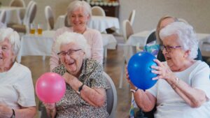 Two family members talking with an elderly loved one and a staff member in a cozy assisted living common area.