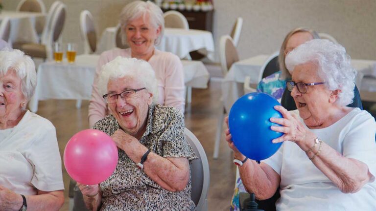 Two family members talking with an elderly loved one and a staff member in a cozy assisted living common area.